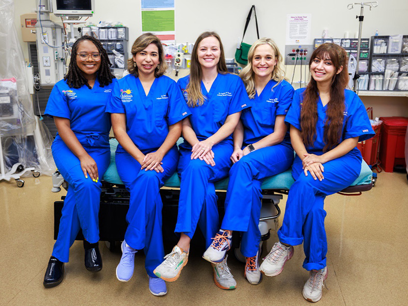 Five pediatric emergency fellows sit together on a gurney in an exam room.